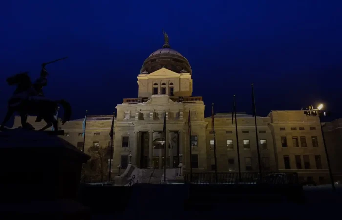 Montana will implement new election management system at a later date 5 The image shows a large, illuminated government building with a central dome at night in Montana, with a silhouetted equestrian statue in the foreground against a deep blue sky.