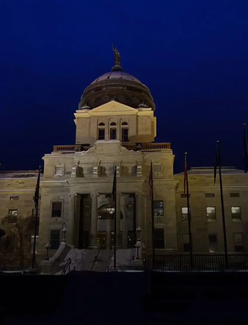 The image shows a large, illuminated government building with a central dome at night in Montana, with a silhouetted equestrian statue in the foreground against a deep blue sky.