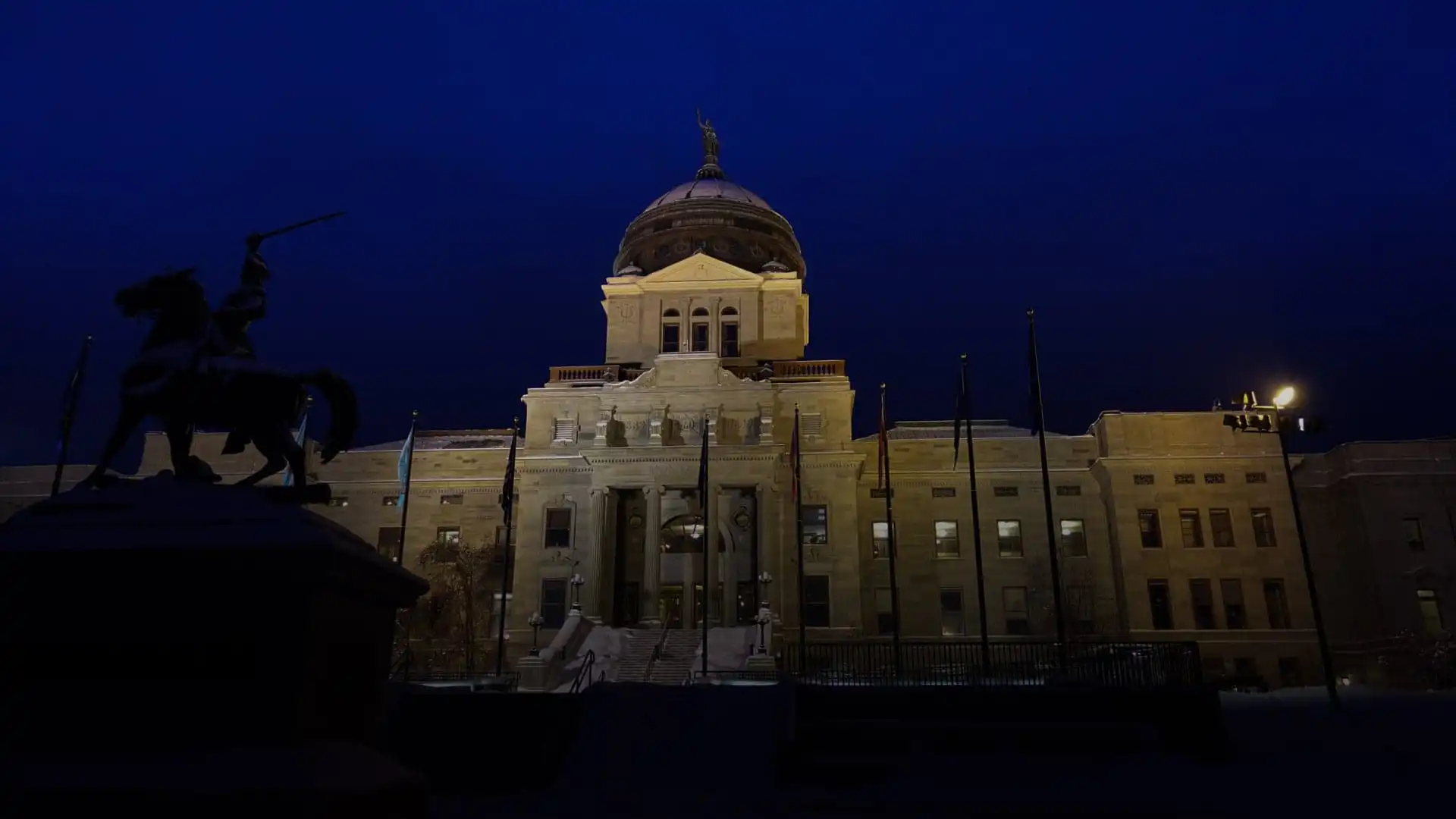 Montana will implement new election management system at a later date 2 The image shows a large, illuminated government building with a central dome at night in Montana, with a silhouetted equestrian statue in the foreground against a deep blue sky.