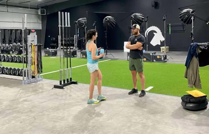 Secretary of State Christi Jacobsen tours MTNTOUGH Fitness Lab; tests grueling sample workout program 6 A woman and a man stand talking in a modern gym with artificial turf, weight racks, and various fitness equipment. The man is wearing shorts and a cap; the woman has on a tank top and shorts.