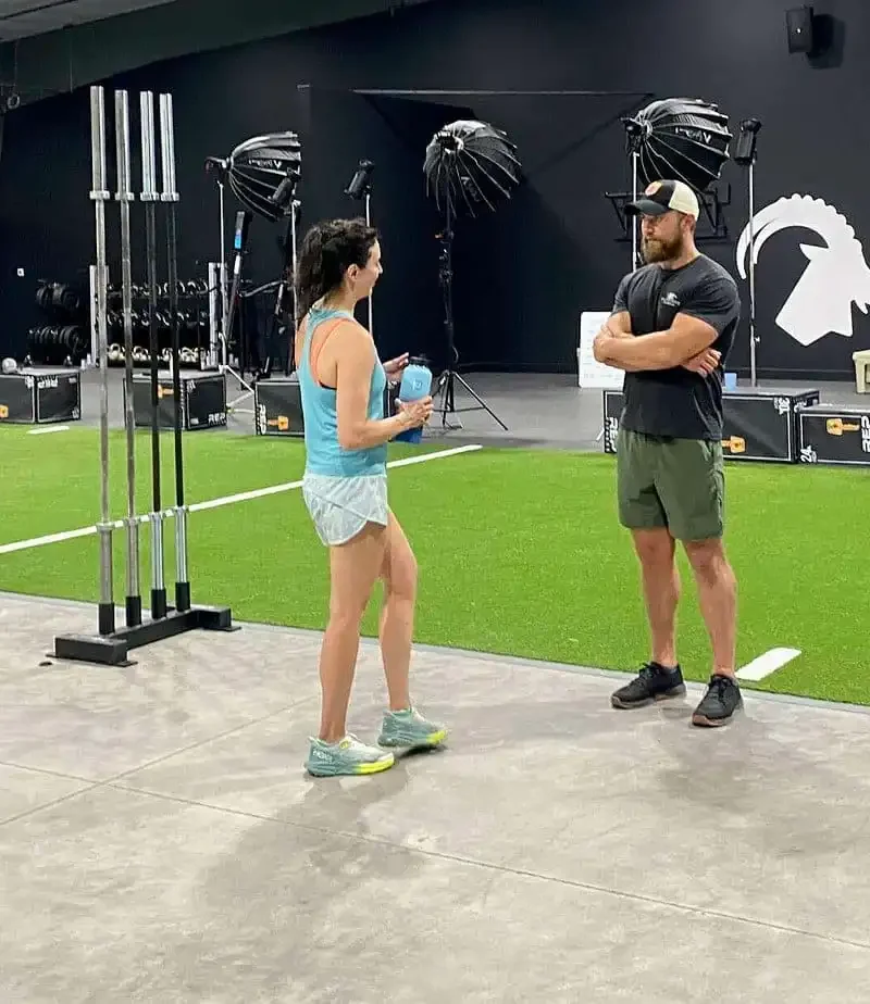 A woman and a man stand talking in a modern gym with artificial turf, weight racks, and various fitness equipment. The man is wearing shorts and a cap; the woman has on a tank top and shorts.