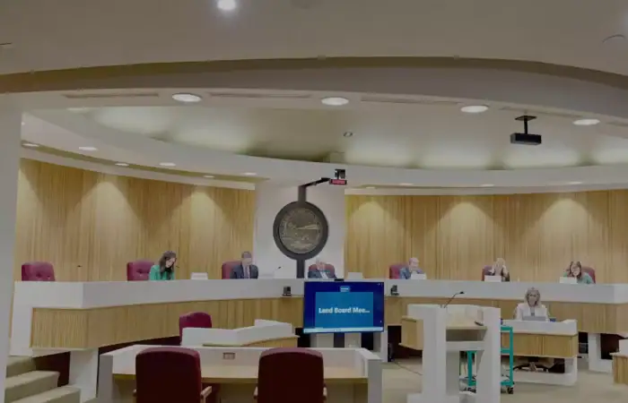 A meeting takes place in a modern government chamber with six people seated at a curved desk, a large seal on the wall, and a screen displaying “Land Board Mee…”—discussing an oil and gas lease valued at $4.5 million.
