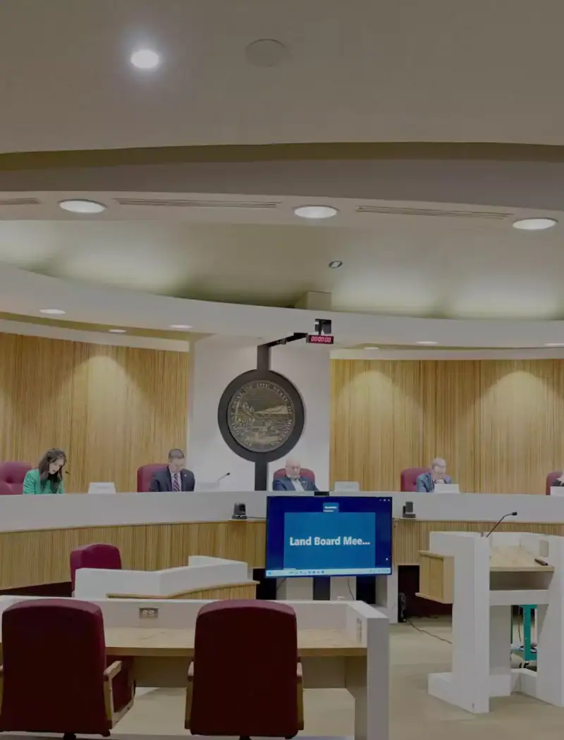 A meeting takes place in a modern government chamber with six people seated at a curved desk, a large seal on the wall, and a screen displaying “Land Board Mee…”—discussing an oil and gas lease valued at $4.5 million.