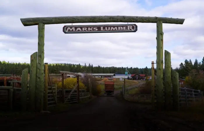 Secretary Jacobsen visits Marks Lumber's 5th annual open house and mill tour 3 A large wooden entrance gate with a sign reading “MARKS LUMBER” stands at the entrance to a rural lumber yard, inviting visitors to explore stacks of wood and trees during an open house mill tour under a cloudy sky.