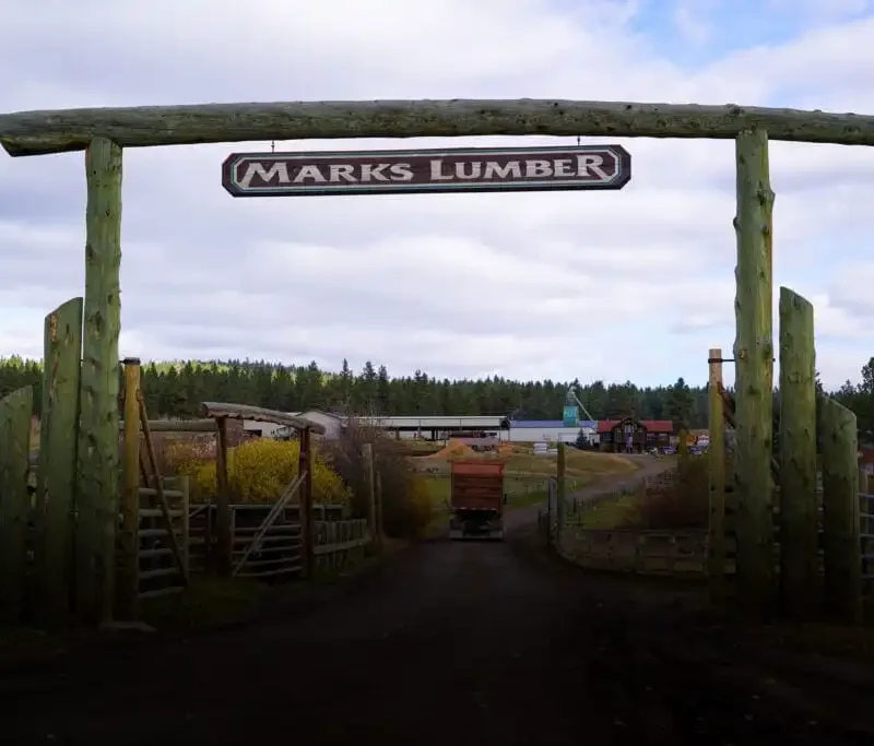 Secretary Jacobsen visits Marks Lumber's 5th annual open house and mill tour 1 A large wooden entrance gate with a sign reading “MARKS LUMBER” stands at the entrance to a rural lumber yard, inviting visitors to explore stacks of wood and trees during an open house mill tour under a cloudy sky.