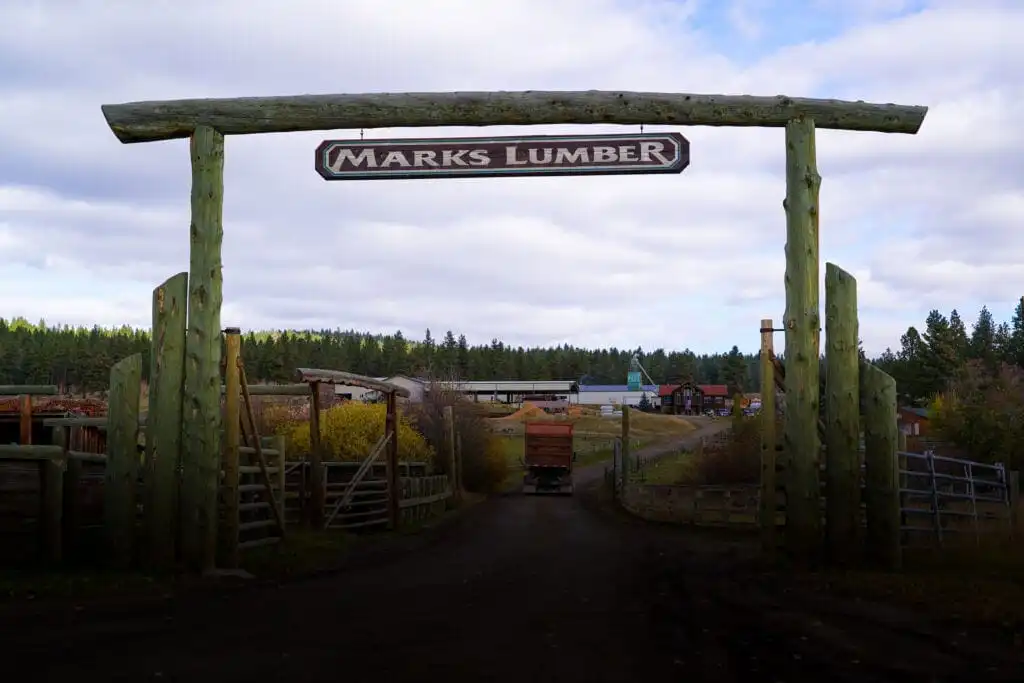 A large wooden entrance gate with a sign reading “MARKS LUMBER” stands at the entrance to a rural lumber yard, inviting visitors to explore stacks of wood and trees during an open house mill tour under a cloudy sky.
