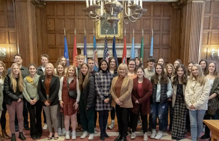 Secretary Christi Jacobsen visits with Great Falls Leadership High School Program 2 A group of students and adults pose together in a wood-paneled room with chandeliers, flags, and a painting in the background. Everyone is standing and smiling for the camera.