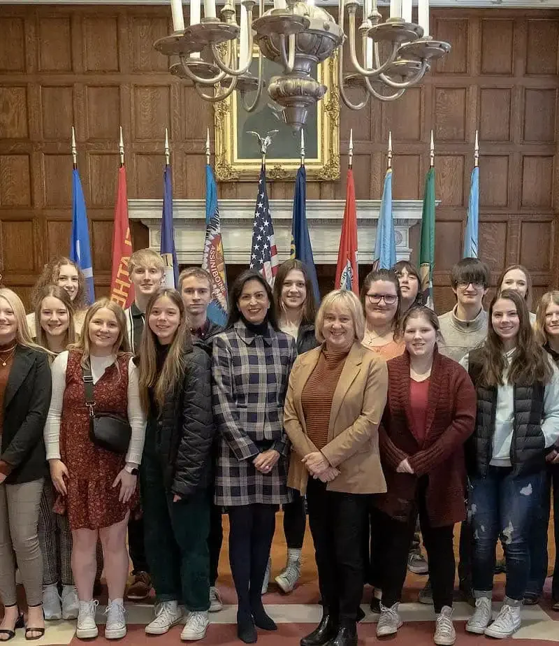 A group of students and adults pose together in a wood-paneled room with chandeliers, flags, and a painting in the background. Everyone is standing and smiling for the camera.