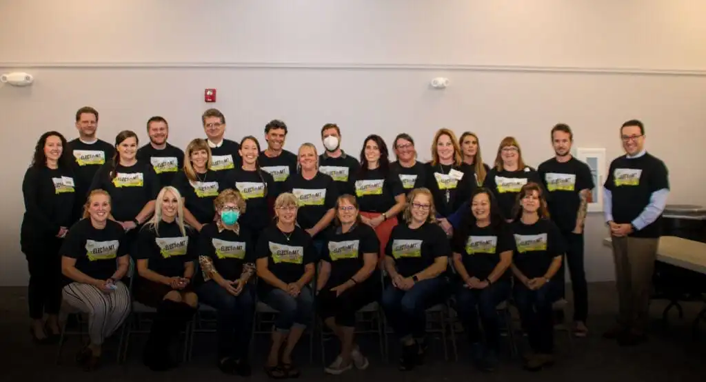 A group of about 25 adults from the Elections Division pose indoors, mostly standing with some seated, all in matching black shirts with a rectangular design. One person in the middle wears a medical mask. They appear to be in a well-lit room.