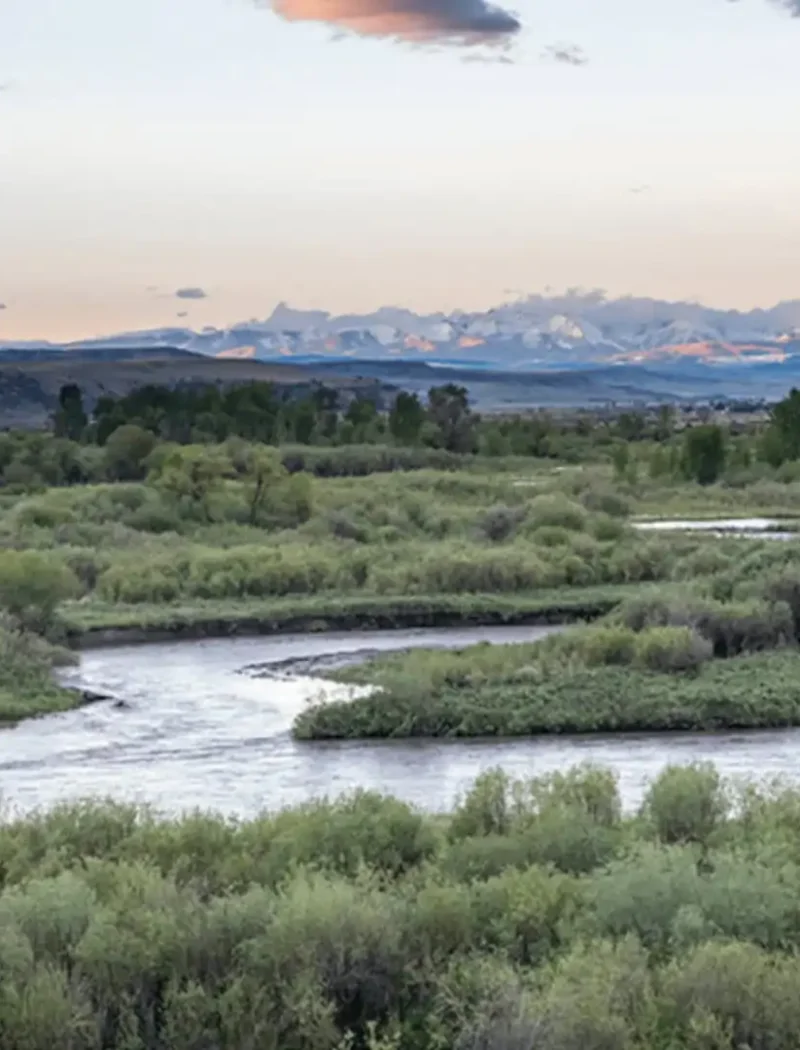 A winding river flows through lush green vegetation at Missouri Headwaters State Park, with distant mountains under a pastel-colored sunset sky—an idyllic landscape preserved through thoughtful acquisition. Clouds are scattered above the scenic view.