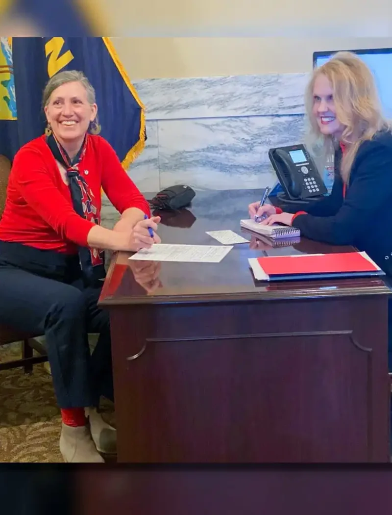 Two women sit across from each other at a wooden office desk, smiling, as papers related to the Montana 2022 candidate filing are spread out. A phone, monitor, flag, and marble wall set the backdrop in this election-focused scene.