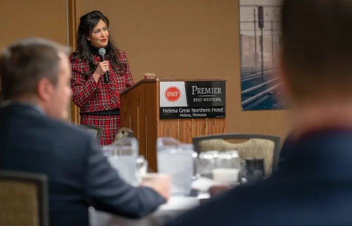 Secretary Christi Jacobsen fights for Montana businesses and forces deceptive mailings to cease 4 A woman in a red plaid outfit speaks at a podium with a microphone in the Best Western Premier Helena Great Northern Hotel, addressing Montana businesses about deceptive mailings. Secretary Christi Jacobsen’s sign is visible, with blurred audience members in the foreground.