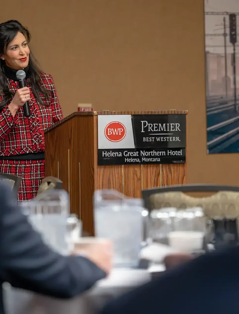 A woman in a red plaid outfit speaks at a podium with a microphone in the Best Western Premier Helena Great Northern Hotel, addressing Montana businesses about deceptive mailings. Secretary Christi Jacobsen’s sign is visible, with blurred audience members in the foreground.