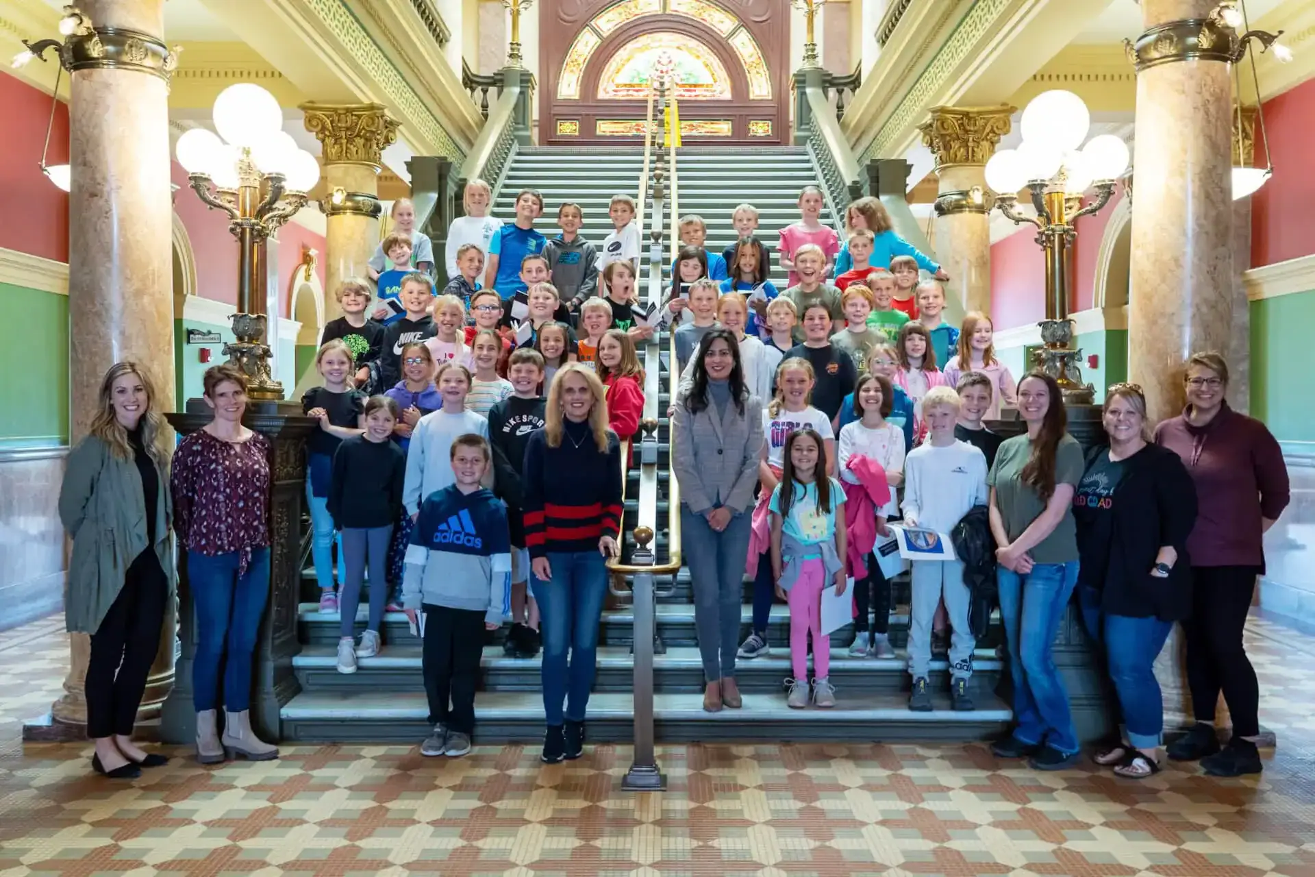 A large group of children and adults pose for a photo on a grand staircase inside a historic building with ornate columns, stained glass, and decorative lighting, celebrating Civics engagement through the YouthVote initiative.