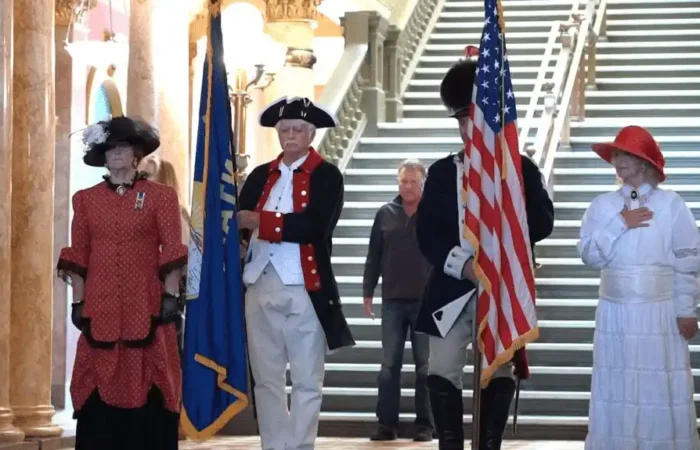 Secretary Jacobsen, Superintendent Arntzen unveil new civics, YouthVote website on Constitution Day 9 Four people in historical costumes stand indoors, two holding flags. Marking Constitution Day, a woman in red and another in white place hands on their chests. A grand staircase and ornate columns are behind them as a casually-dressed man looks on.