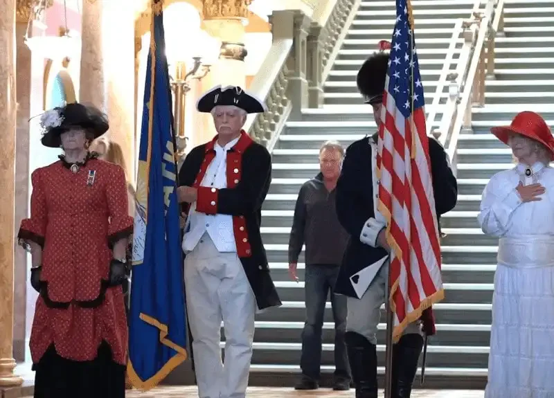 Secretary Jacobsen, Superintendent Arntzen unveil new civics, YouthVote website on Constitution Day 10 Four people in historical costumes stand indoors, two holding flags. Marking Constitution Day, a woman in red and another in white place hands on their chests. A grand staircase and ornate columns are behind them as a casually-dressed man looks on.