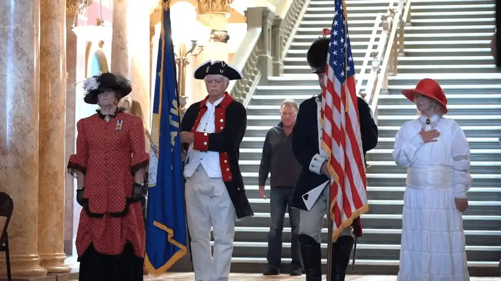 Four people in historical costumes stand indoors, two holding flags. Marking Constitution Day, a woman in red and another in white place hands on their chests. A grand staircase and ornate columns are behind them as a casually-dressed man looks on.