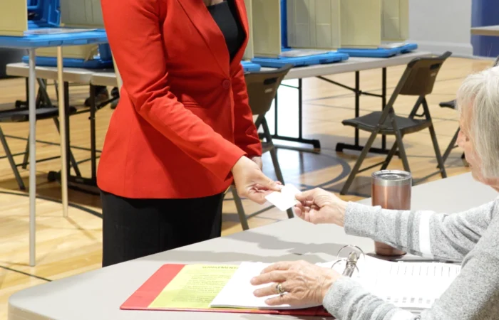 'Victory for the integrity of elections': Secretary Christi Jacobsen celebrates court decision on Montana's voter ID law 2 A woman in a red blazer hands an ID card to a seated poll worker at a voting station with booths and chairs in the background, reflecting procedures put in place under the Montana voter ID law to ensure election integrity.