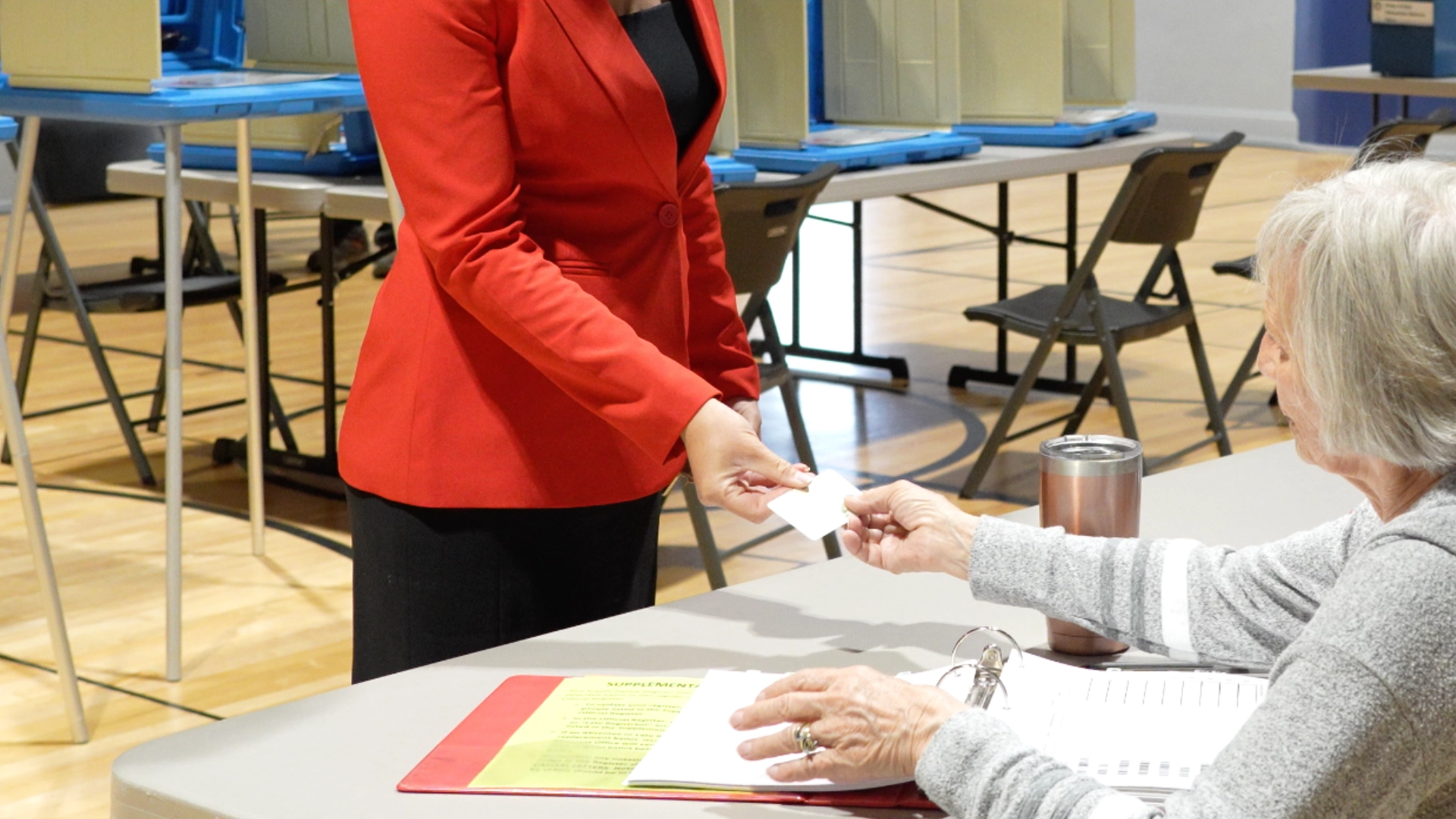 A woman in a red blazer hands an ID card to a seated poll worker at a voting station with booths and chairs in the background, reflecting procedures put in place under the Montana voter ID law to ensure election integrity.