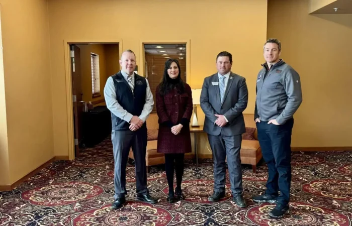 Secretary Christi Jacobsen champions business growth and civic engagement at Montana Professional Firefighters Legislative Convention 1 Four people stand side by side in a room with tan walls and patterned carpet at the Montana Professional Firefighters Legislative Convention. Three men in business attire and a woman in a dark coat pose, highlighting civic engagement. Two armchairs are in the background.