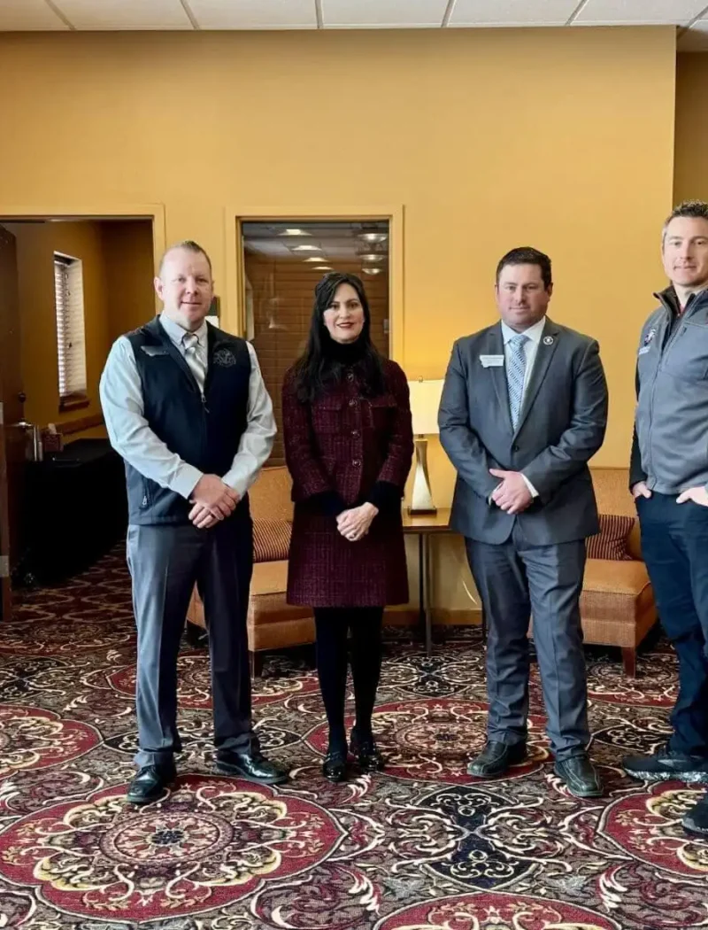 Four people stand side by side in a room with tan walls and patterned carpet at the Montana Professional Firefighters Legislative Convention. Three men in business attire and a woman in a dark coat pose, highlighting civic engagement. Two armchairs are in the background.