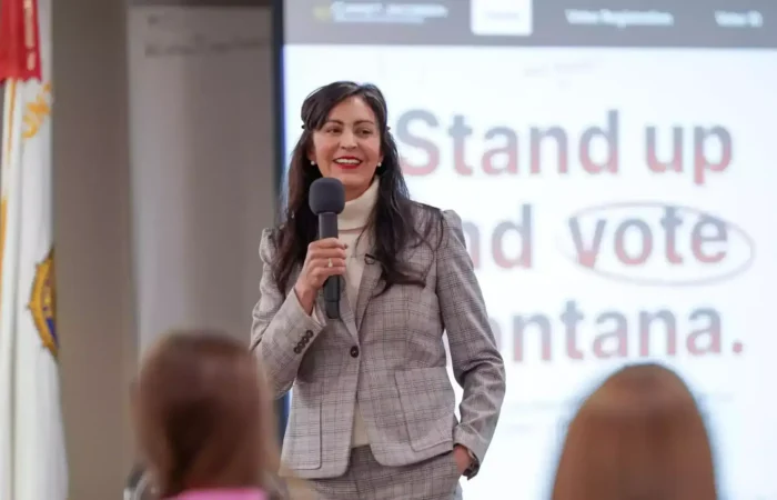 Secretary Christi Jacobsen: 'Montana Elections Team celebrates National Voter Registration Month' 4 A woman in a gray plaid suit, possibly Montana Elections’ Christi Jacobsen, holds a microphone and smiles while speaking to an audience. Behind her, a screen displays Stand up and vote, highlighting National Voter Registration Month.