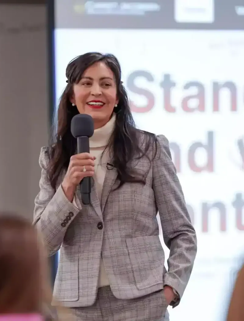 A woman in a gray plaid suit, possibly Montana Elections’ Christi Jacobsen, holds a microphone and smiles while speaking to an audience. Behind her, a screen displays Stand up and vote, highlighting National Voter Registration Month.