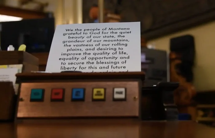 A close-up of a wooden panel with colored buttons labeled, in front of a card displaying part of Montana’s constitution about liberty, opportunity, and equality—highlighting bipartisan efforts in the Montana Senate. The background is out of focus.
