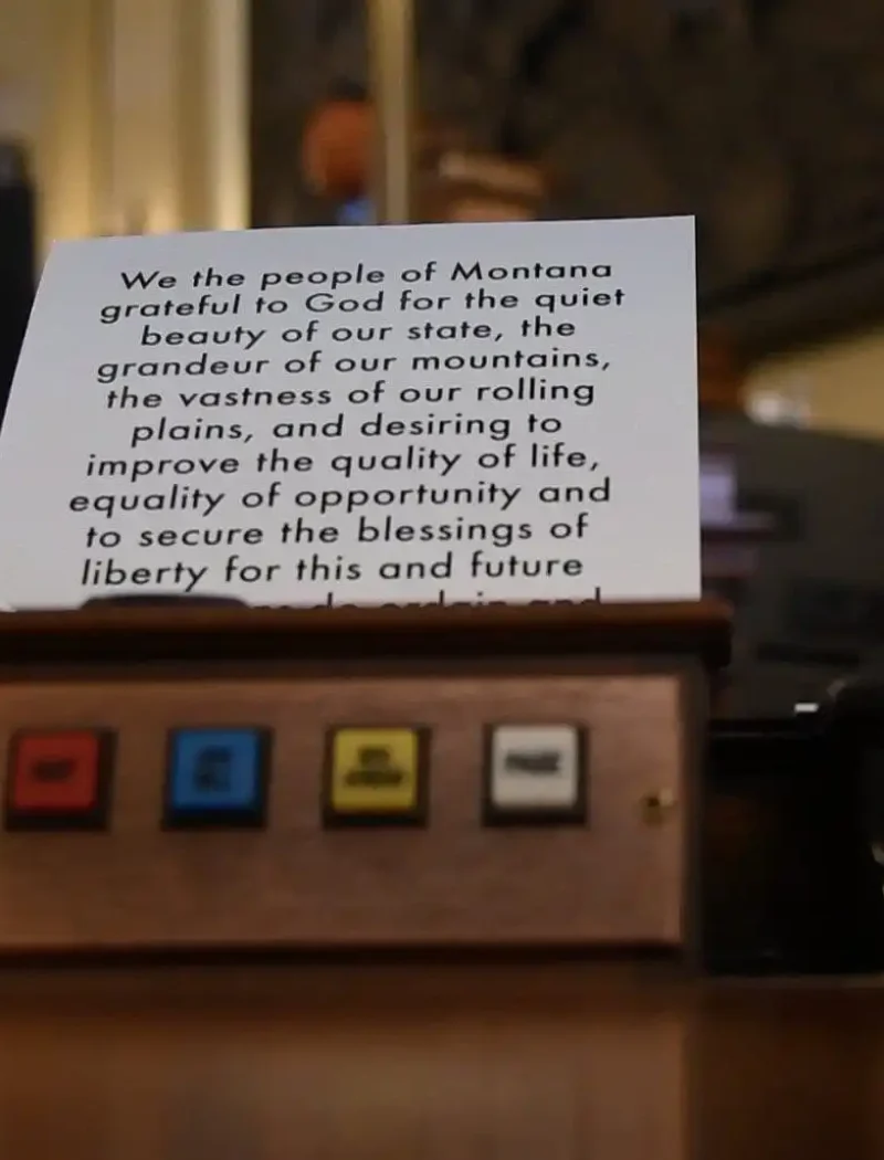 A close-up of a wooden panel with colored buttons labeled, in front of a card displaying part of Montana’s constitution about liberty, opportunity, and equality—highlighting bipartisan efforts in the Montana Senate. The background is out of focus.