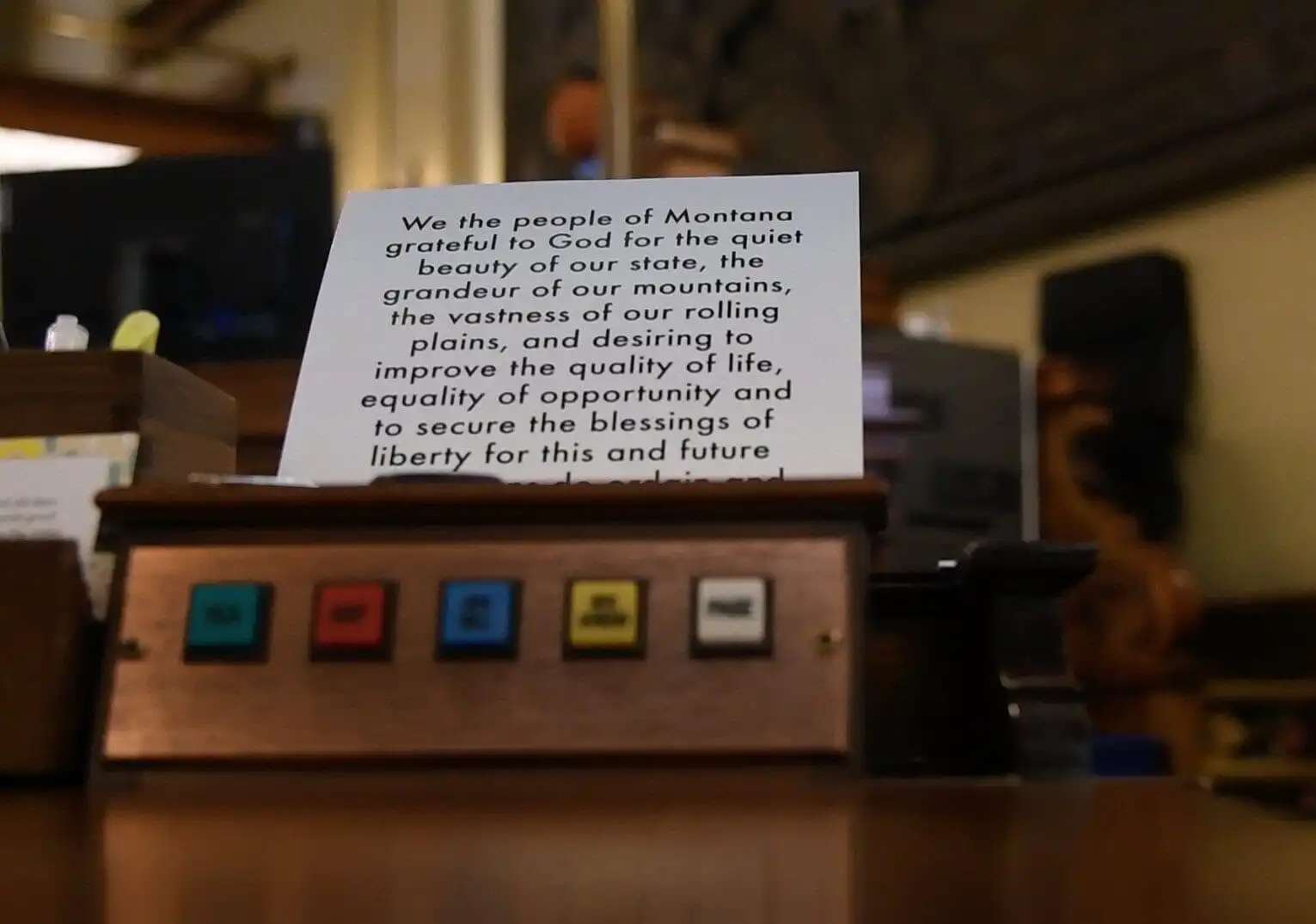 A close-up of a wooden panel with colored buttons labeled, in front of a card displaying part of Montana’s constitution about liberty, opportunity, and equality—highlighting bipartisan efforts in the Montana Senate. The background is out of focus.
