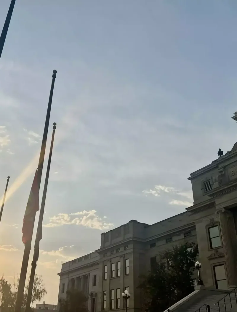 The sun sets behind Montana’s government building with a dome, as several flags fly at half-staff in the warm evening light of August 2024. The sky is partly cloudy, hinting at opportunities for new businesses on the horizon.