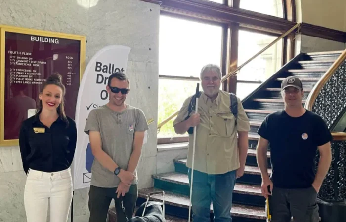 History Made: Secretary Jacobsen celebrates 'colossal victory' for Montana's blind & visually impaired voters 2 Four people stand together indoors near a staircase. Two men, Montana blind voters, use mobility canes—one with a guide dog. Secretary Jacobsen, in business attire, stands on the left. Behind them is a directory board and a “Ballot Drop Off” sign.