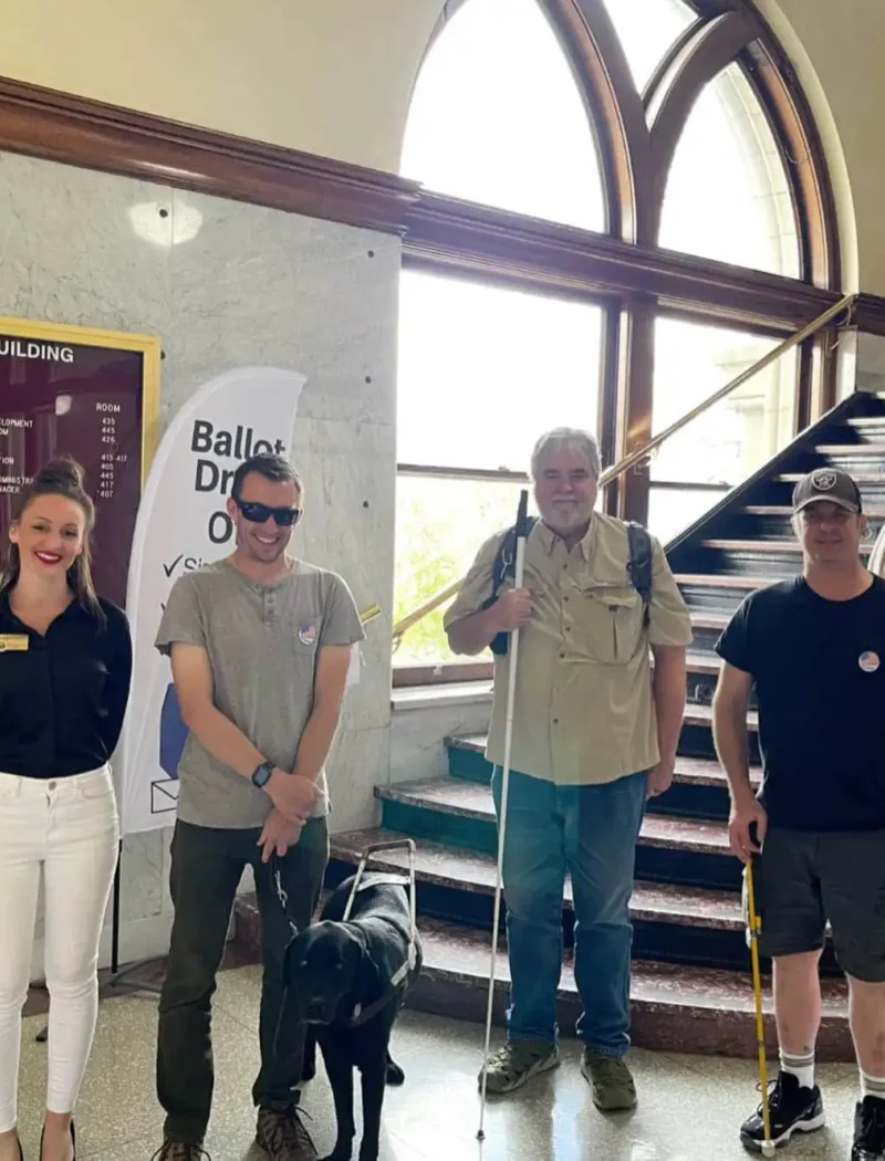 Four people stand together indoors near a staircase. Two men, Montana blind voters, use mobility canes—one with a guide dog. Secretary Jacobsen, in business attire, stands on the left. Behind them is a directory board and a “Ballot Drop Off” sign.