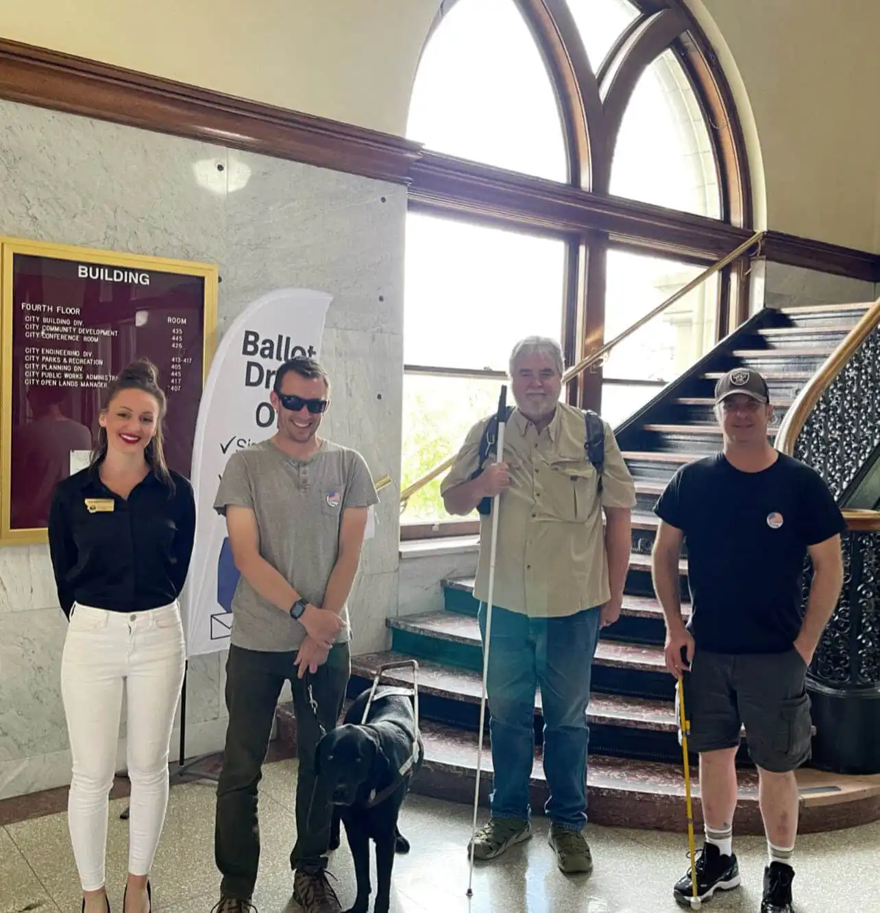 Four people stand together indoors near a staircase. Two men, Montana blind voters, use mobility canes—one with a guide dog. Secretary Jacobsen, in business attire, stands on the left. Behind them is a directory board and a “Ballot Drop Off” sign.