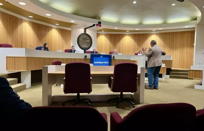 A man stands and presents to a panel of officials seated at a curved desk in a council chamber. Empty chairs face the front. A large circular emblem and digital screen display Land Board 11/21 in Montana’s Treasure State.