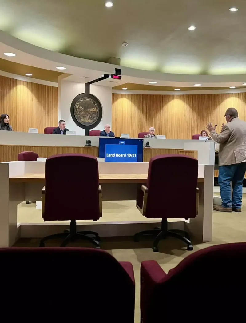 A man stands and presents to a panel of officials seated at a curved desk in a council chamber. Empty chairs face the front. A large circular emblem and digital screen display Land Board 11/21 in Montana’s Treasure State.