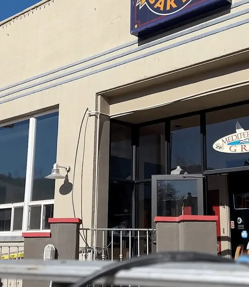 A beige building with signs reading Park Avenue Bakery & Cafe and Mediterranean Grill above a glass door. The entrance has a small patio with railings and red accents, perfect for enjoying Earl Grey Cake or fresh French bread under the blue sky.