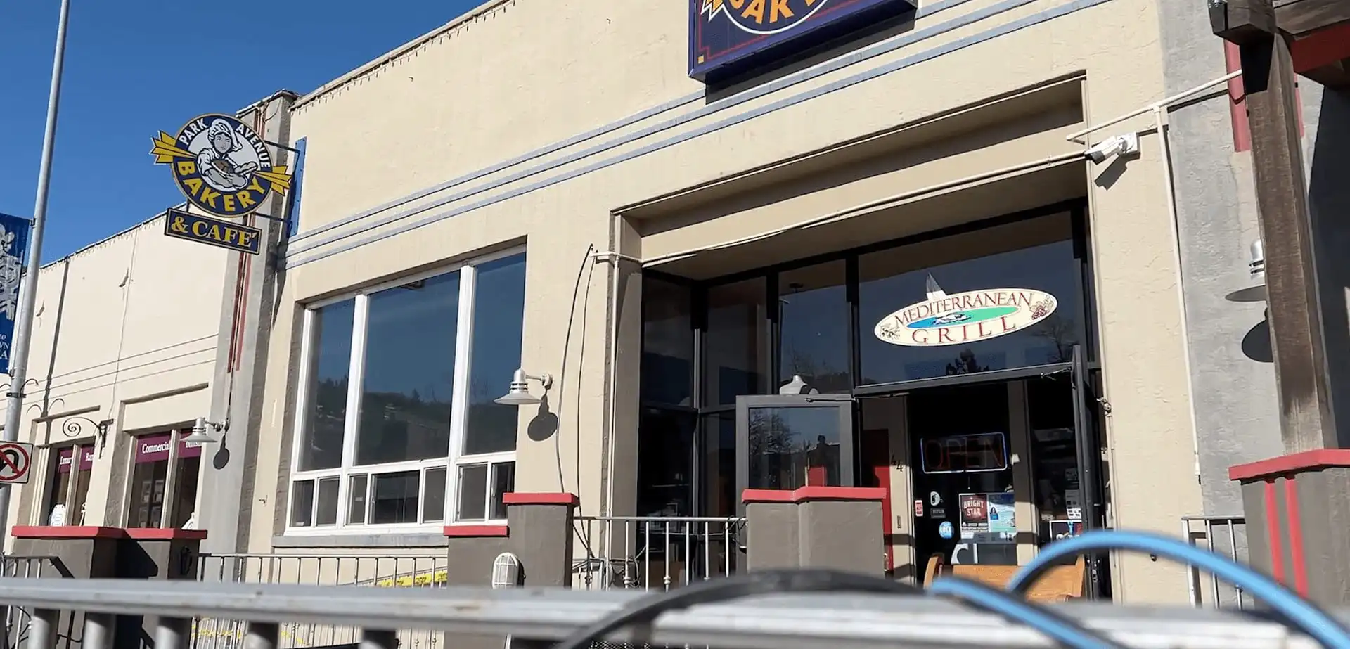 A beige building with signs reading Park Avenue Bakery & Cafe and Mediterranean Grill above a glass door. The entrance has a small patio with railings and red accents, perfect for enjoying Earl Grey Cake or fresh French bread under the blue sky.