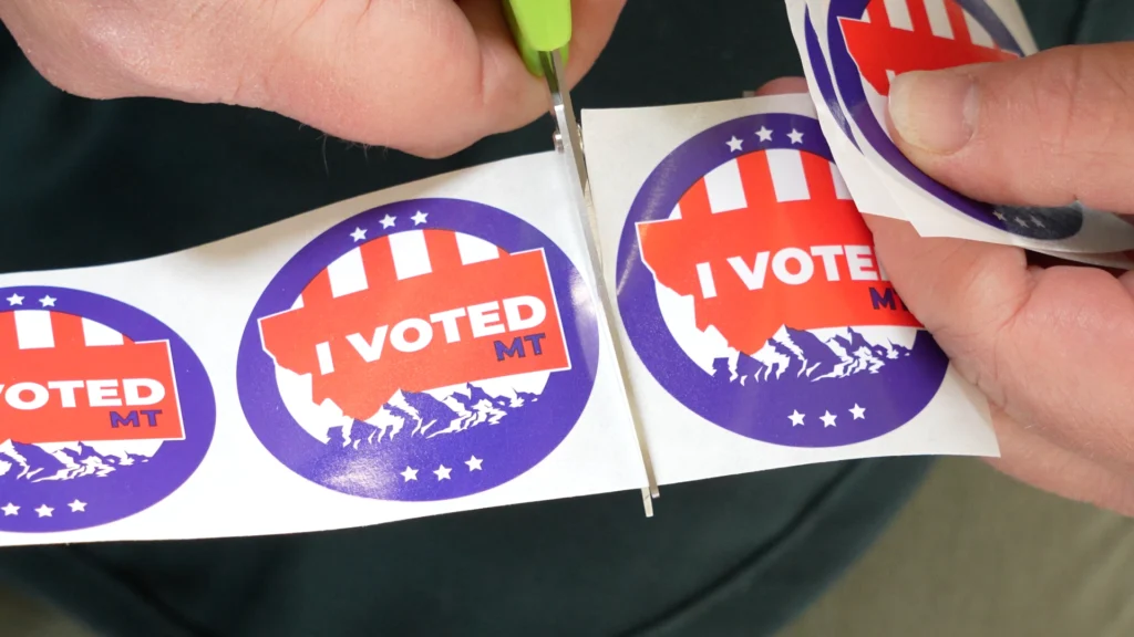 Christi Jacobsen - Montana Secretary of State 9 A person uses scissors to cut out a round I VOTED MT sticker from a strip of similar stickers, each with a patriotic design and the outline of Montana—a reminder of the vital role poll worker recruitment plays in supporting elections, especially on National Poll Worker Recruitment Day, led by Secretary Christi Jacobsen.