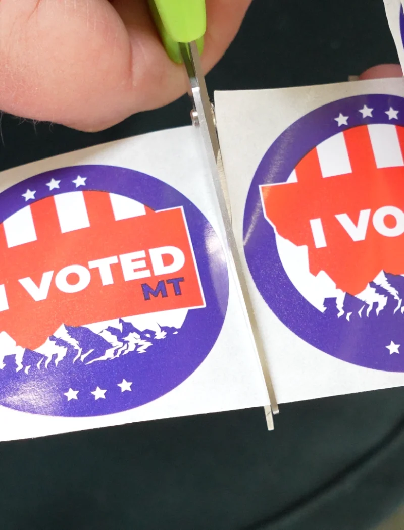 A person uses scissors to cut out a round I VOTED MT sticker from a strip of similar stickers, each with a patriotic design and the outline of Montana—a reminder of the vital role poll worker recruitment plays in supporting elections, especially on National Poll Worker Recruitment Day, led by Secretary Christi Jacobsen.