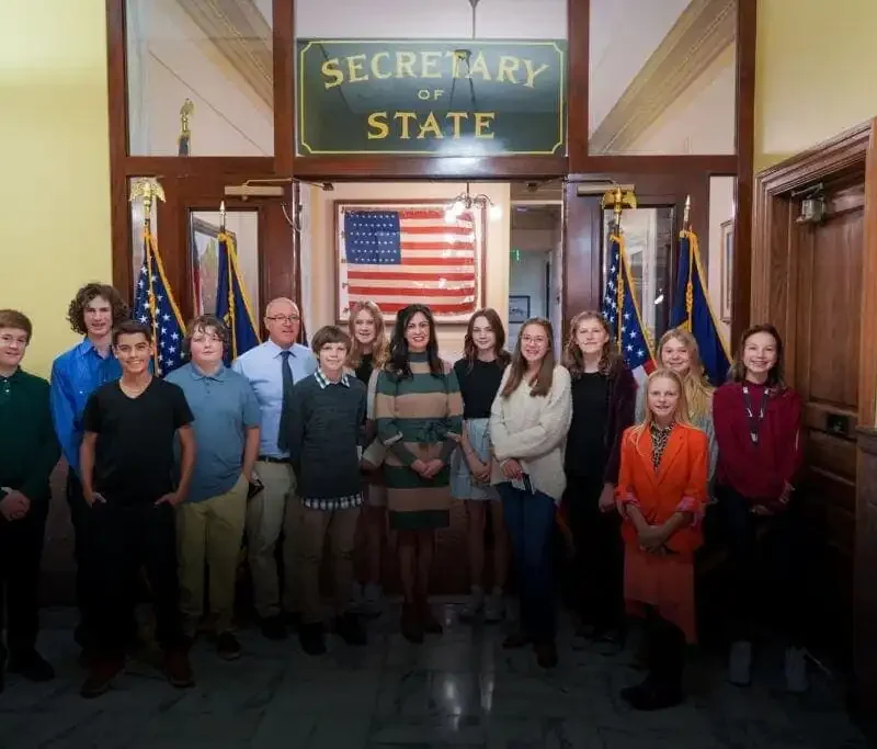 A group from Roosevelt Middle School, including student council members and adults, pose for a group photo in front of a Secretary of States Office sign and several American flags inside a government building.