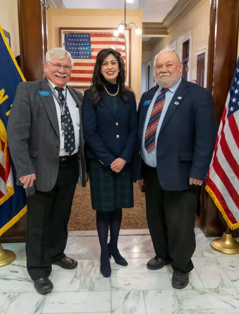 Three people stand smiling in a hallway between two pairs of U.S. and Montana flags, dressed in business attire. An American flag hangs in the background, highlighting the significance of the 2024 election and Montana candidate filing season.