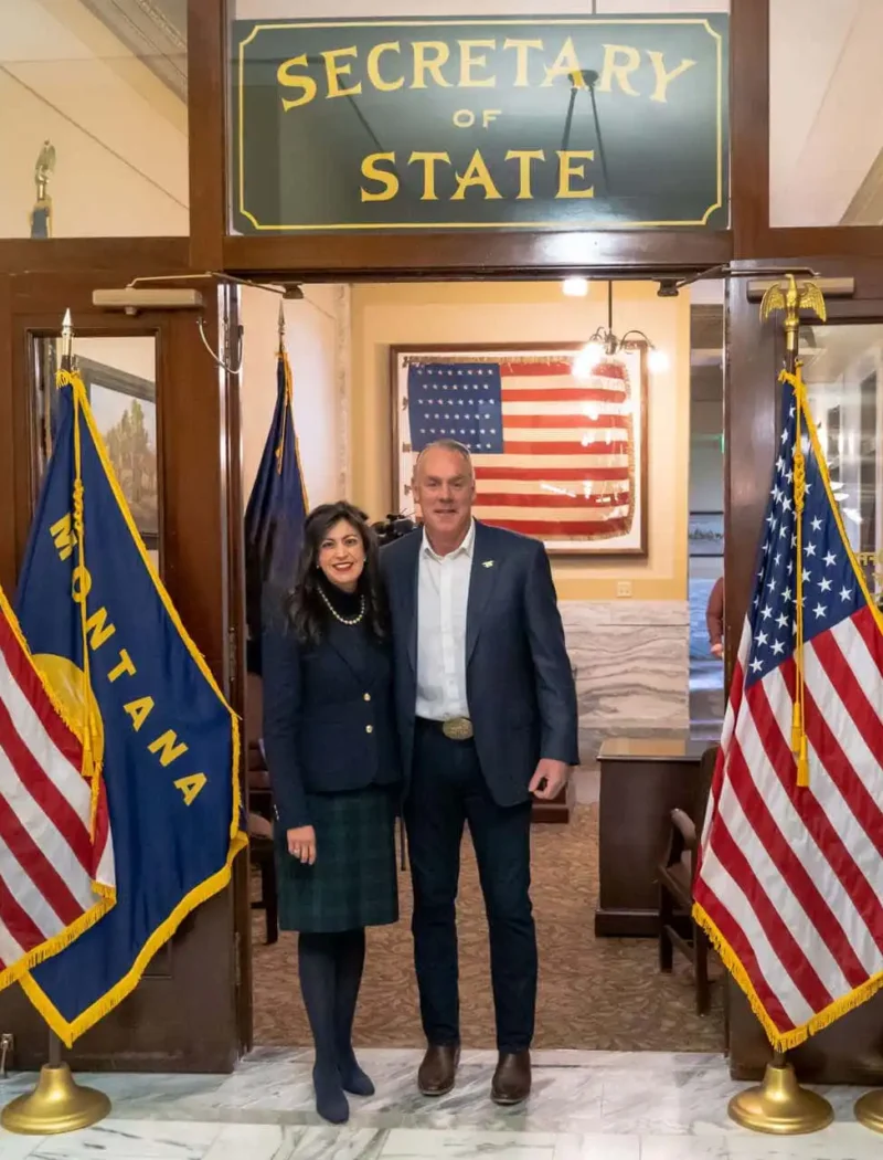 Two people stand smiling under a Secretary of State sign, surrounded by Montana and U.S. flags in an official-looking building with marble floors—possibly celebrating recent candidate filings—with an American flag in the background.