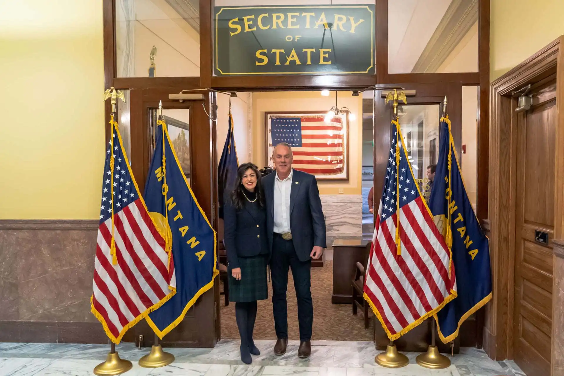 Two people stand smiling under a Secretary of State sign, surrounded by Montana and U.S. flags in an official-looking building with marble floors—possibly celebrating recent candidate filings—with an American flag in the background.