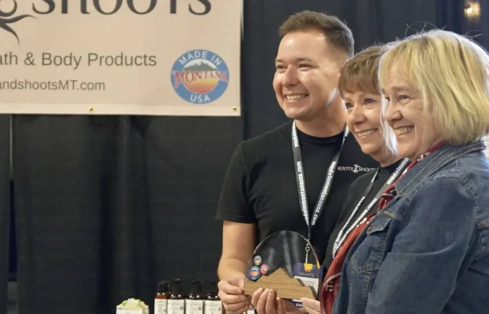 Three people smile and pose for a photo at the 2025 Tradeshow booth for Roots & Shoots handcrafted bath and body products. One person holds an award. Shelves with Made in Montana products and a company sign are visible in the background.