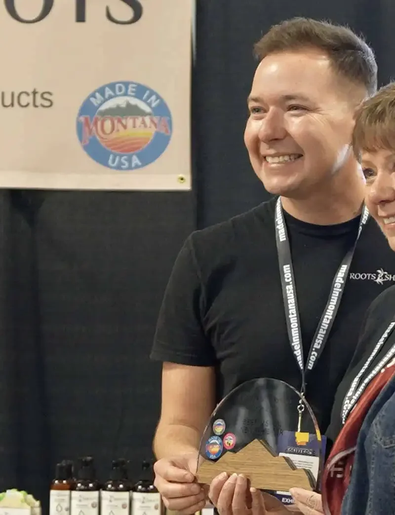 Three people smile and pose for a photo at the 2025 Tradeshow booth for Roots & Shoots handcrafted bath and body products. One person holds an award. Shelves with Made in Montana products and a company sign are visible in the background.