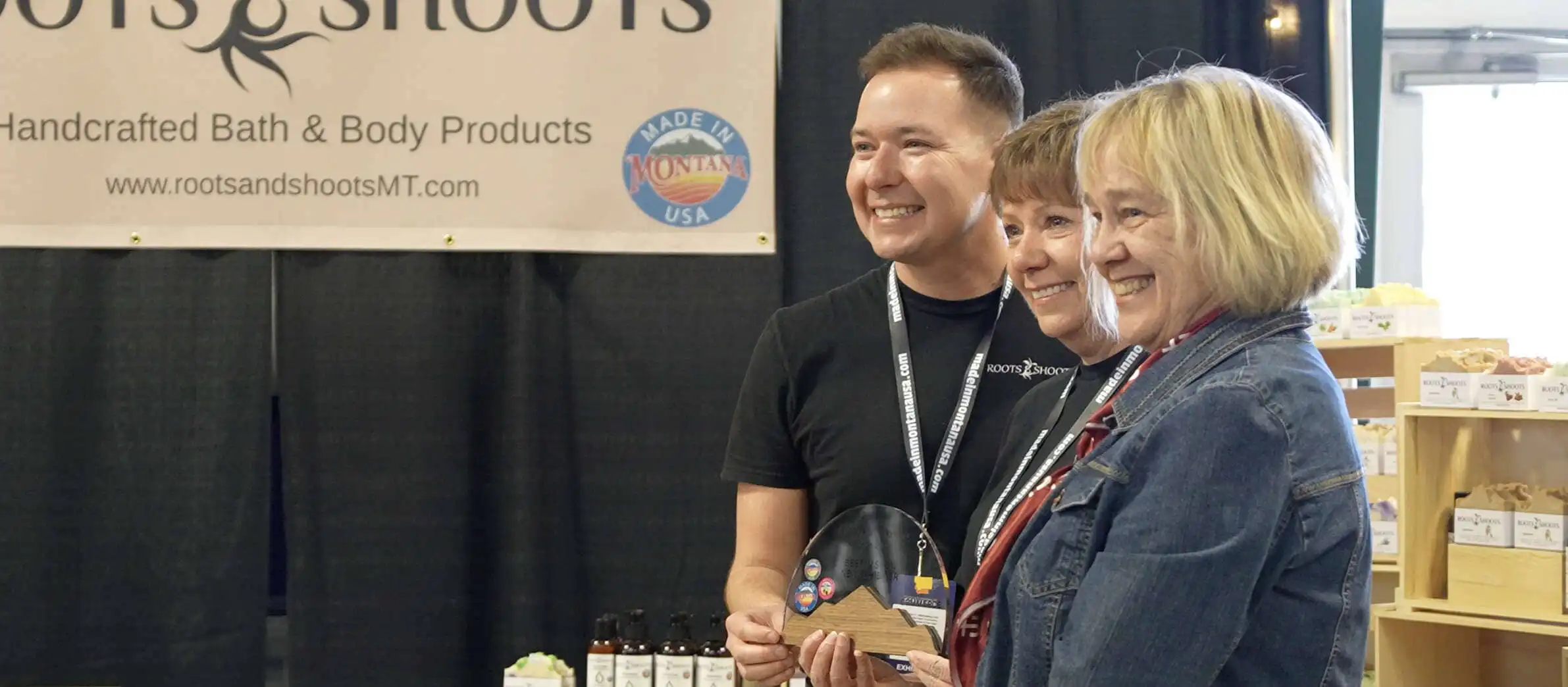 Three people smile and pose for a photo at the 2025 Tradeshow booth for Roots & Shoots handcrafted bath and body products. One person holds an award. Shelves with Made in Montana products and a company sign are visible in the background.