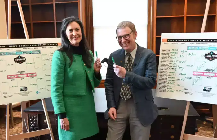Secretary Christi Jacobsen, Commissioner James Brown make NCAA Tournament selections 10 Two people smiling and holding green markers stand in front of two whiteboards filled with handwritten NCAA basketball tournament brackets in an office setting with shelves and a window in the background.