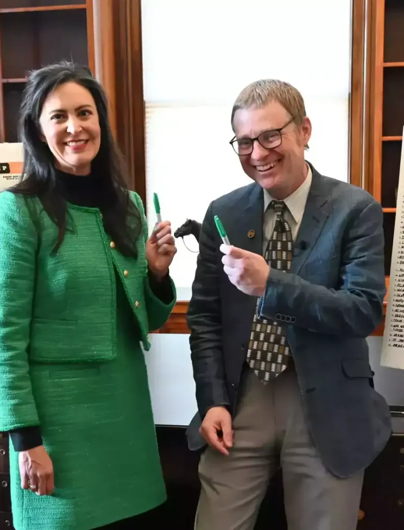 Two people smiling and holding green markers stand in front of two whiteboards filled with handwritten NCAA basketball tournament brackets in an office setting with shelves and a window in the background.