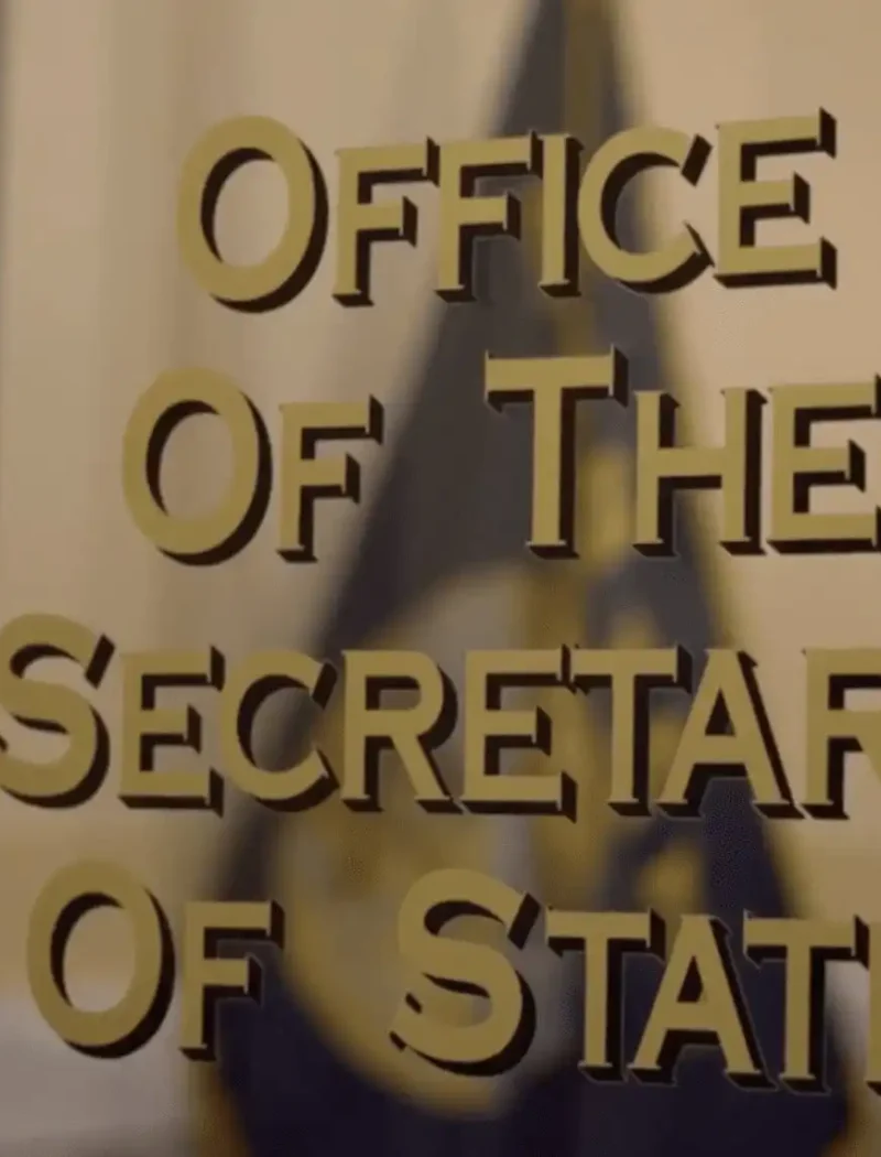 Glass door with gold lettering that reads Office Of The Secretary Of State, referencing Montana elections, with a blurred flag and interior visible in the background.